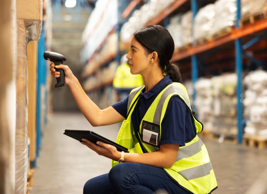 Trabalhadora de logística em armazém escaneando etiqueta com um leitor de código de barras e conferindo produtos em prateleiras.