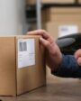 Pessoa usando leitor de código de barras para registrar uma caixa de papelão em ambiente de depósito ou armazém.