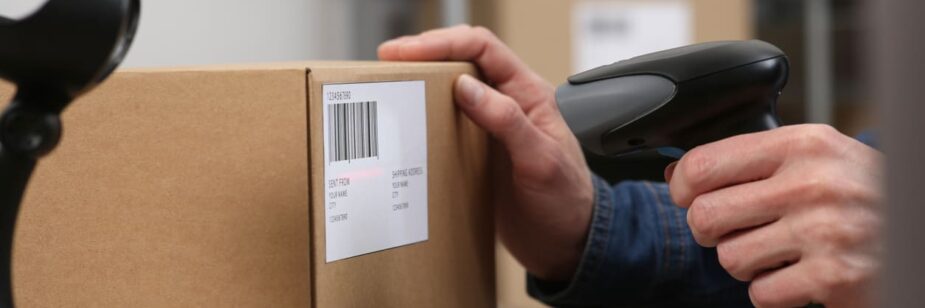 Pessoa usando leitor de código de barras para registrar uma caixa de papelão em ambiente de depósito ou armazém.