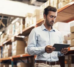 Homem de negócios conferindo inventário em armazém com prateleiras cheias de caixas de papelão, utilizando um tablet para gestão e controle de estoque.