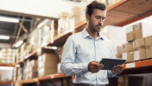 Homem de negócios conferindo inventário em armazém com prateleiras cheias de caixas de papelão, utilizando um tablet para gestão e controle de estoque.