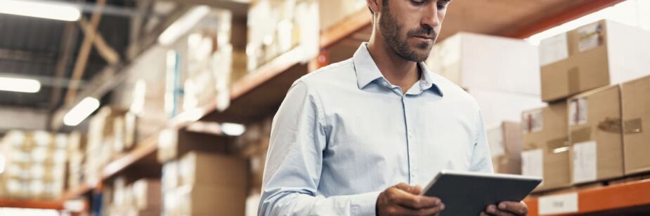 Homem de negócios conferindo inventário em armazém com prateleiras cheias de caixas de papelão, utilizando um tablet para gestão e controle de estoque.