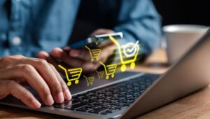 Pessoa utilizando laptop e smartphone para vender online, com ícones de carrinho de compras digital representando comércio eletrônico e compras na internet.