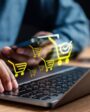 Pessoa utilizando laptop e smartphone para vender online, com ícones de carrinho de compras digital representando comércio eletrônico e compras na internet.