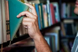 Pessoa organizando e guardando um livro em uma estante, com a capa verde sendo puxada para trás entre livros em uma biblioteca, ao lado de outras prateleiras.