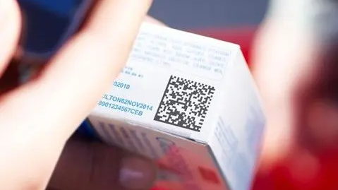 Pessoa segurando um medicamento com código QR impresso, em primeiro plano, com fundo desfocado.