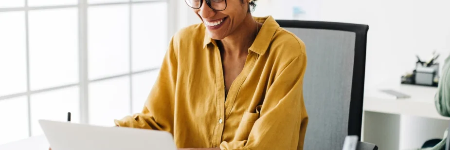 Mulher sorridente com óculos trabalha em escritório moderno, usando notebook sobre a mesa, enquanto olha a tela com expressão de satisfação.