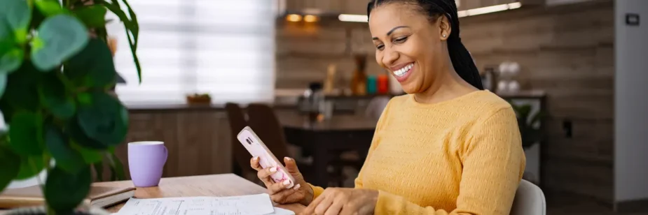 Mulher sorridente em casa usa o celular enquanto trabalha à mesa com documentos e pastas na cozinha moderna, com planta ao lado e ambiente iluminado