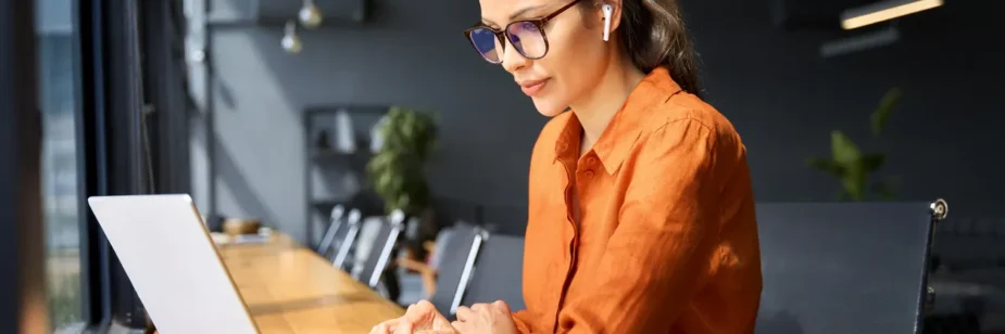 Mulher em um escritório moderno usando óculos e trabalhando em um notebook na mesa de madeira, com tela e ambiente corporativo ao fundo.