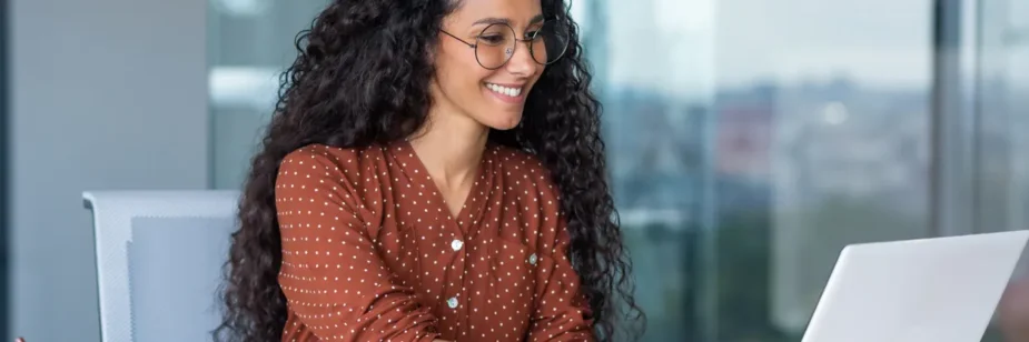Mulher com cabelo cacheado e óculos usa notebook no escritório, sorrindo enquanto trabalha à mesa com documentos e caneta sobre o computador.